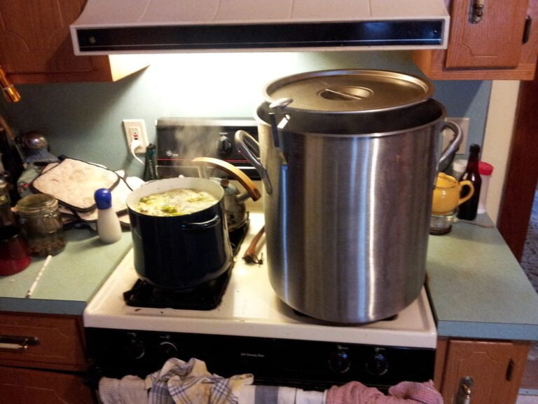 A large stainless steel pot with a lid and a smaller black pot with boiling contents on a stove in a kitchen with various cooking utensils and items around.
