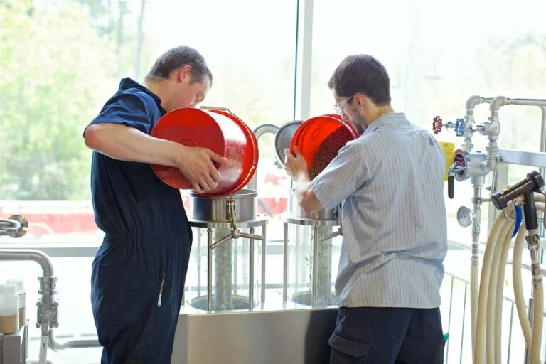 Two men pouring ingredients into a commercial brewery tank with brewing equipment in the background.
