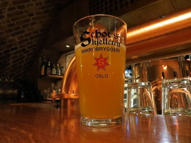 A pint of hazy beer in a branded glass on a wooden bar counter with empty glasses in the background and dim lighting reflecting off metal surfaces.