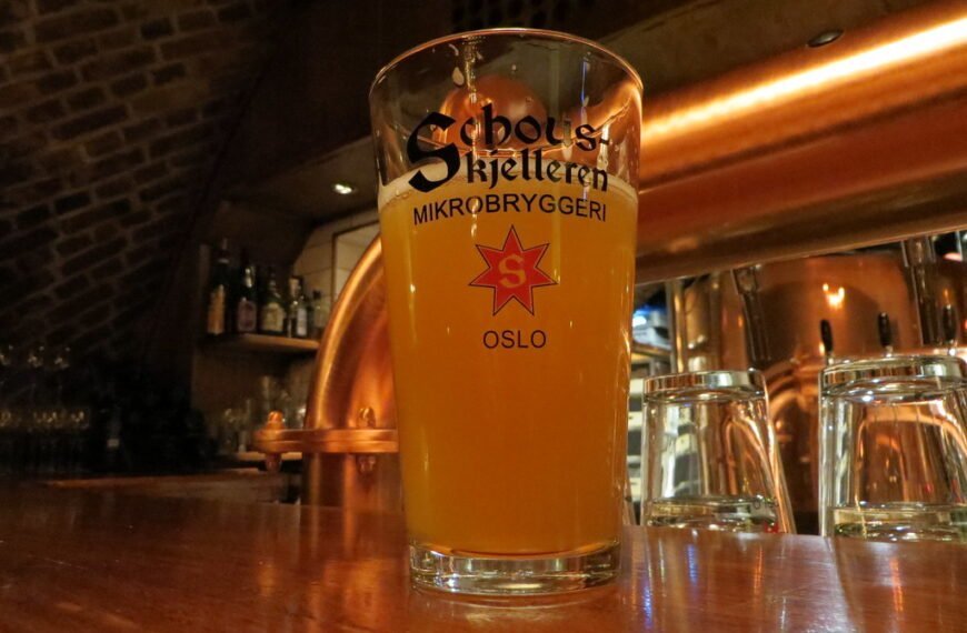 A pint of hazy beer in a branded glass on a wooden bar counter with empty glasses in the background and dim lighting reflecting off metal surfaces.