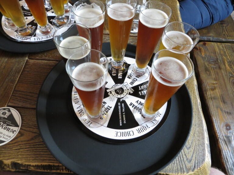 A flight of various beers in tall glasses arranged on a round tasting board on a wooden table.