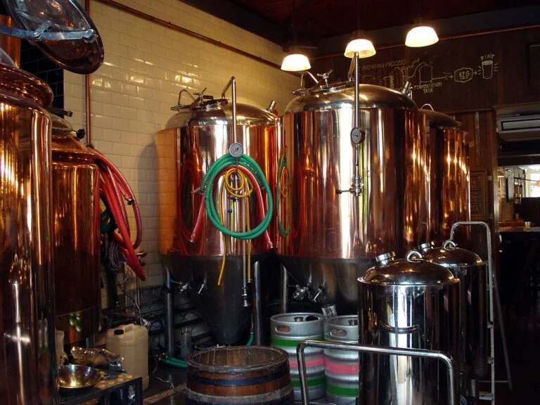 Brewery interior with shiny copper brewing vats, hoses, and brewing equipment, with a diagram of the brewing process on the wall in the background.