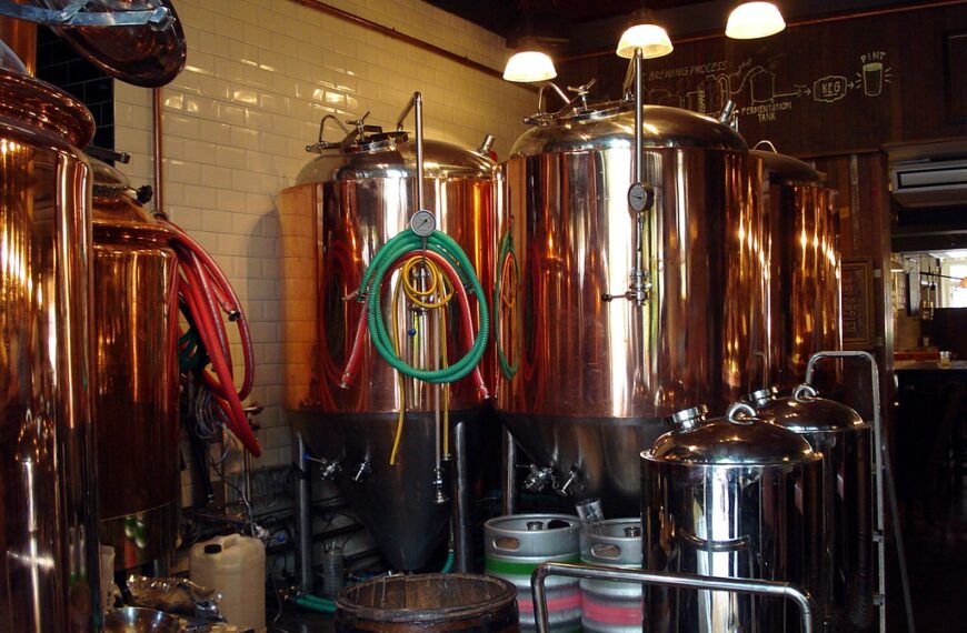 Brewery interior with shiny copper brewing vats, hoses, and brewing equipment, with a diagram of the brewing process on the wall in the background.