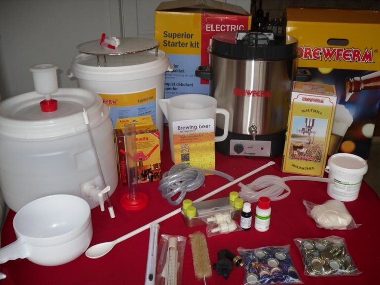 Home beer brewing kit with various equipment, including fermenting buckets, a malt mill, a brewing kettle, hoses, measuring instruments, and ingredients on a red tablecloth.