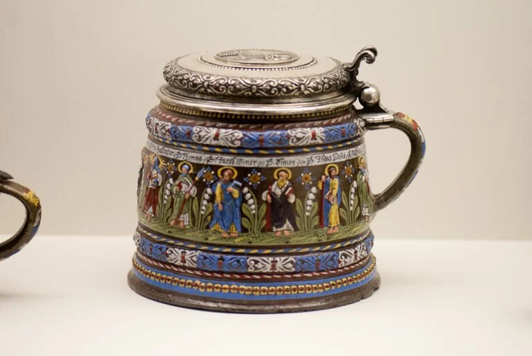 Decorative beer stein with a pewter lid featuring religious figures in medieval attire painted around the circumference, set against a white background.