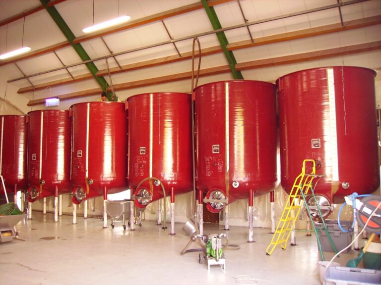 Interior of a winery with a row of large red fermentation tanks, winemaking equipment on the floor, and fluorescent lights overhead.
