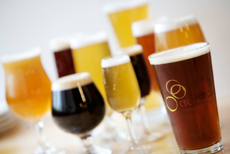 A variety of beers in different hues, served in glasses arranged on a table, with the focus on a pint of amber beer with the "CBC Cambridge Brewing Co." logo.