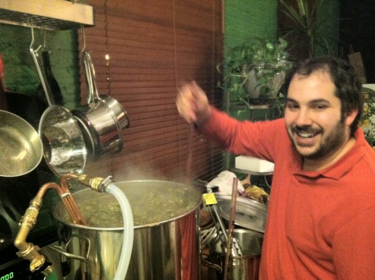 A smiling person stirring a large pot with brewing equipment around in a home brewing setup.