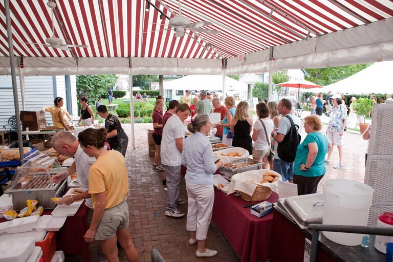 People standing in line under a large red and white striped tent at an outdoor food market with vendors serving from stalls.