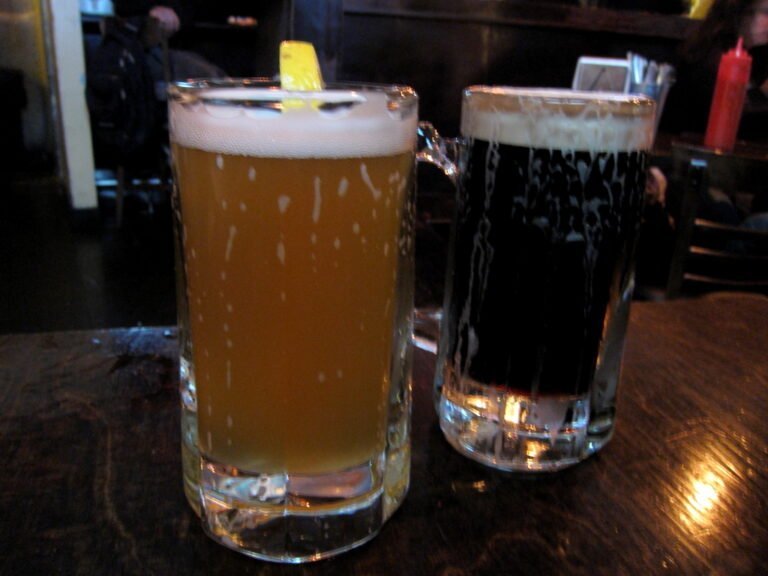 Two glasses of beer on a wooden table, one light-colored and hazy, and the other dark, possibly a stout, both with a frothy head.