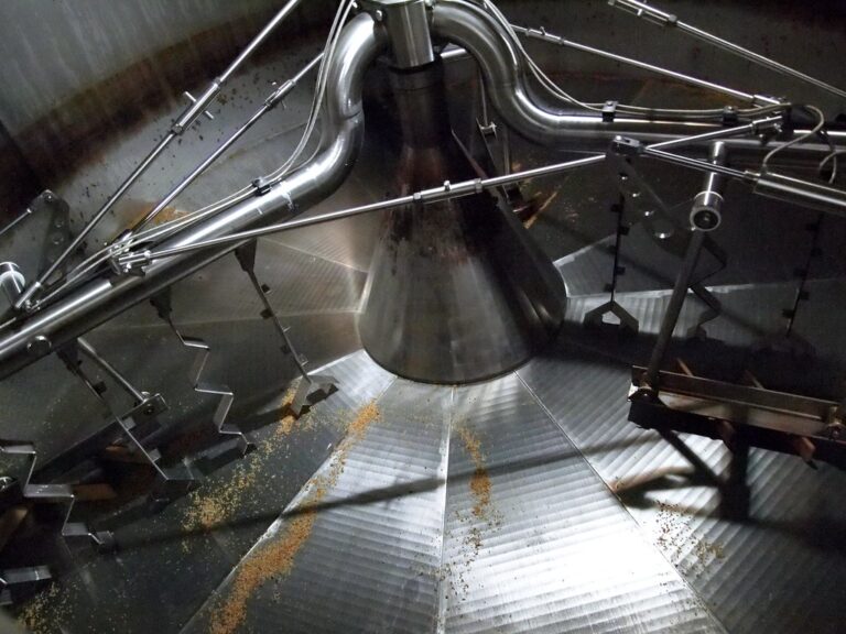 Interior view of an industrial brewing vat showing the stirring mechanism and scattered hops on the ribbed metal floor.