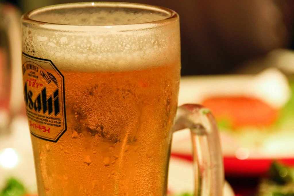 A close-up of a cold Asahi beer in a glass with condensation and frothy head, with a blurred background of a dining setting.