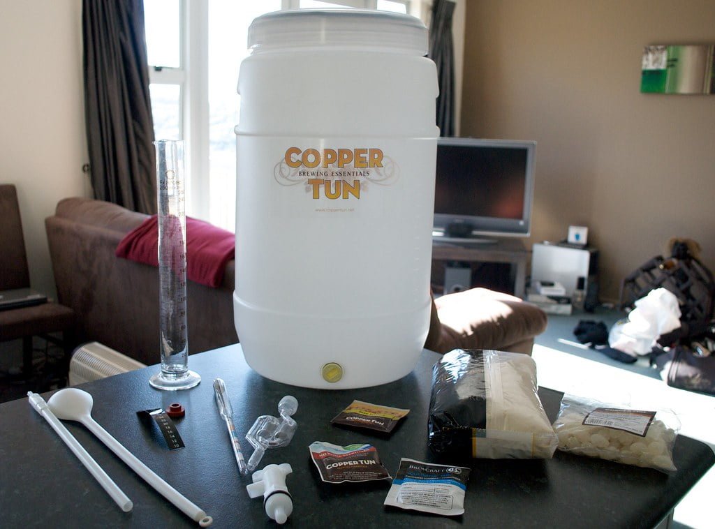 Homebrewing equipment and ingredients on a table including a fermentation bucket, hydrometer, various spoons, a thermometer, yeast packets, and bags of hops and grains, with a living room background.