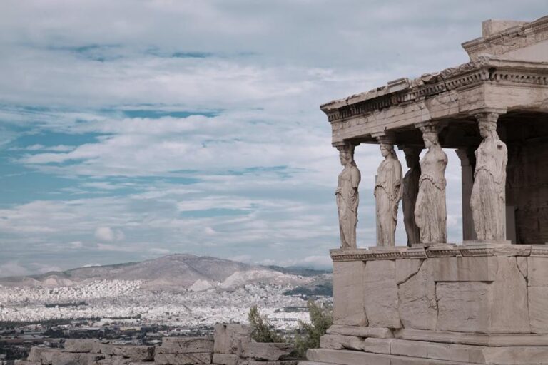 The Caryatids on the Erechtheion at the Acropolis of Athens, with a backdrop of a cloudy sky and the cityscape in the distance.