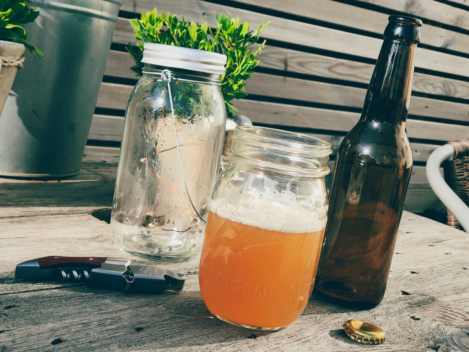 A mason jar filled with a frothy orange beverage, an empty glass bottle, and a brown beer bottle on a wooden table, accompanied by a folded pocketknife and a bottle cap. In the background, potted plants adorn the scene.