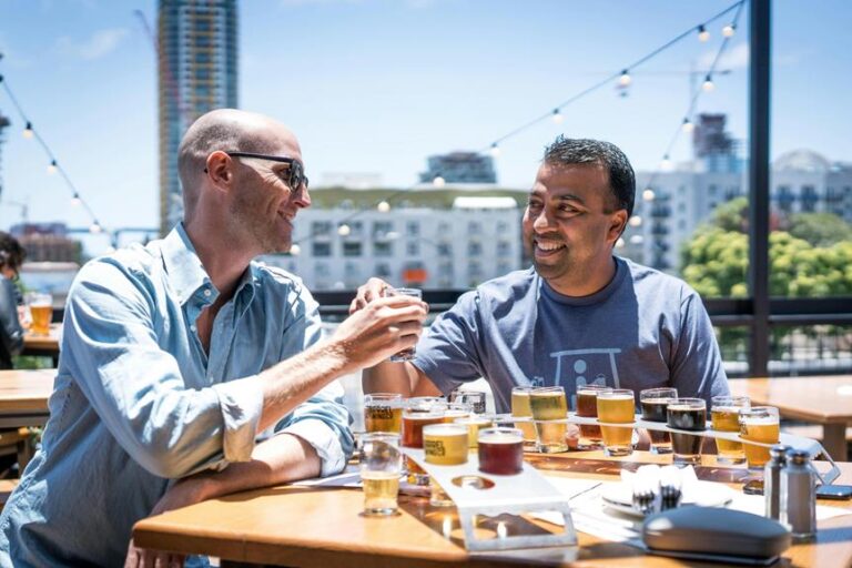 Two men are toasting with beers at an outdoor brewery with various glasses of beer on the table and a cityscape in the background.