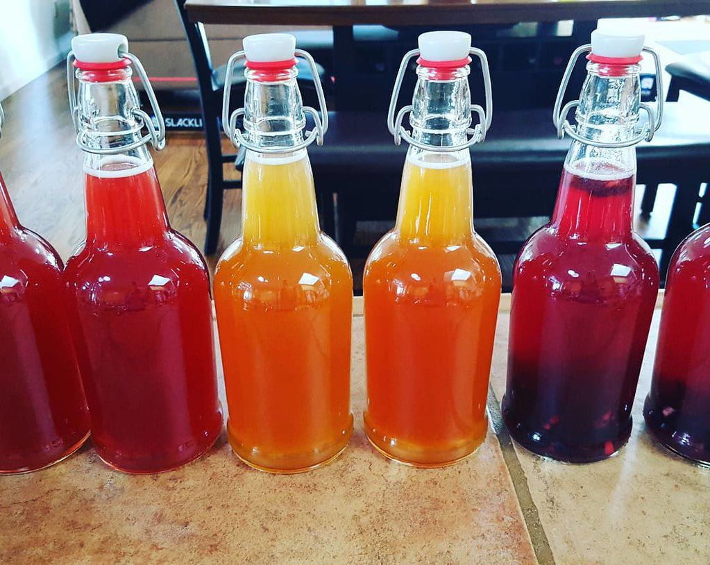 Seven glass bottles with swing-top closures containing liquids in shades of red and orange, arranged in a line on a table.