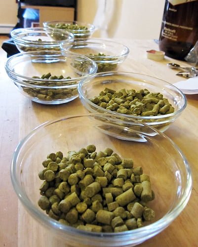 A line of glass bowls on a wooden surface, each containing a different quantity of green hop pellets, commonly used for brewing beer.