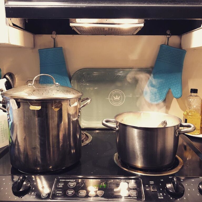 Two pots cooking on a stove with steam rising and a pair of blue silicone oven mitts hanging above.