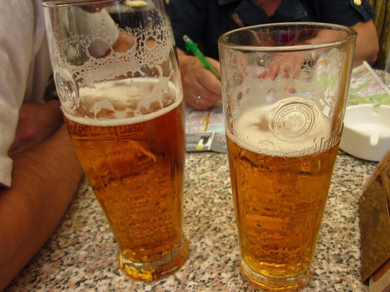 Two partially filled glasses of beer on a table with people in the background holding pens and a map.