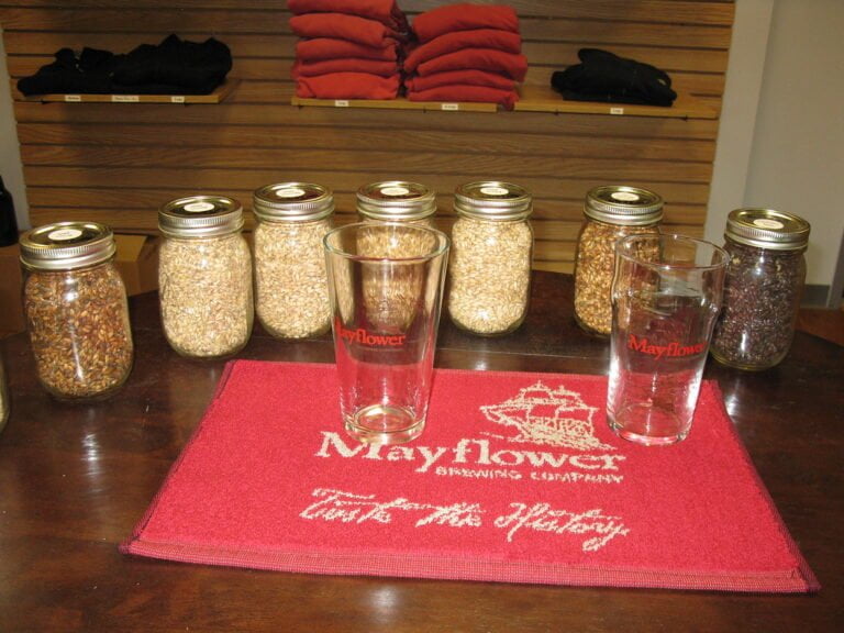 Six mason jars filled with different grains on a wooden counter, two branded pint glasses, and a red mat with "Mayflower Brewing Company" text. Wooden shelving with folded clothing is visible in the background.