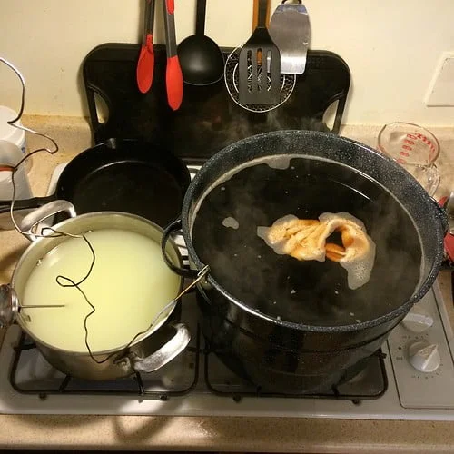Two pots on a stove: one with clear boiling water and a pair of tongs holding a piece of food, the other containing a milky liquid with a wire whisk. Kitchen utensils hang in the background.