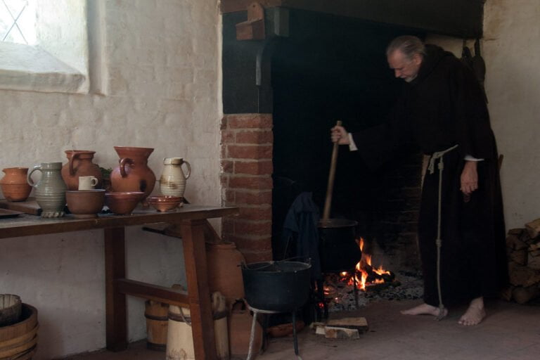A person in a traditional monk's robe stirs a pot over an open fire in a rustic kitchen with various pottery on a wooden table.