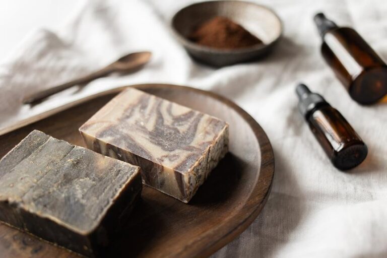 Two bars of homemade soap with a marbled appearance are placed on a wooden tray, next to a wooden spoon and a small bowl of brown powder. In the background, there are two dark amber bottles with droppers, all arranged on a textured white fabric.