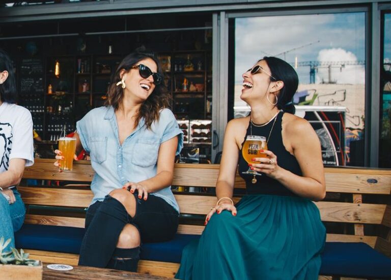 Two women laughing and sitting outdoors on a bench with drinks in their hands, with a bar counter and bottles in the background.