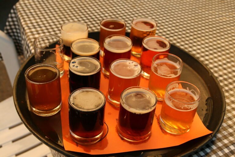 A variety of beer glasses with different colored beers on a round tray, sitting on a black and white checkered tabletop.