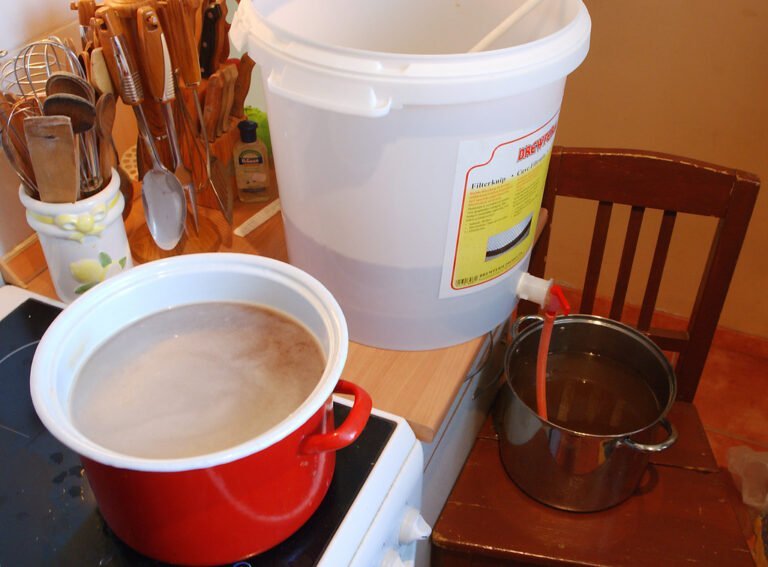 A home brewing setup with a large white fermentation bucket with a spigot above a metal pot on a chair, next to a red enamel pot on a stovetop, and a collection of kitchen utensils in the background.