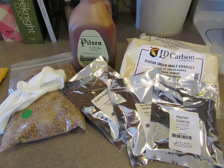 Home brewing ingredients laid out on a kitchen counter, including a plastic jug of Pilsen malt extract, bags of hop pellets labeled Czech Saaz, East Kent Goldings, and Styrian Golding, a packet of Briess dried malt extract, and a plastic bag of grains with a small white cloth bag.