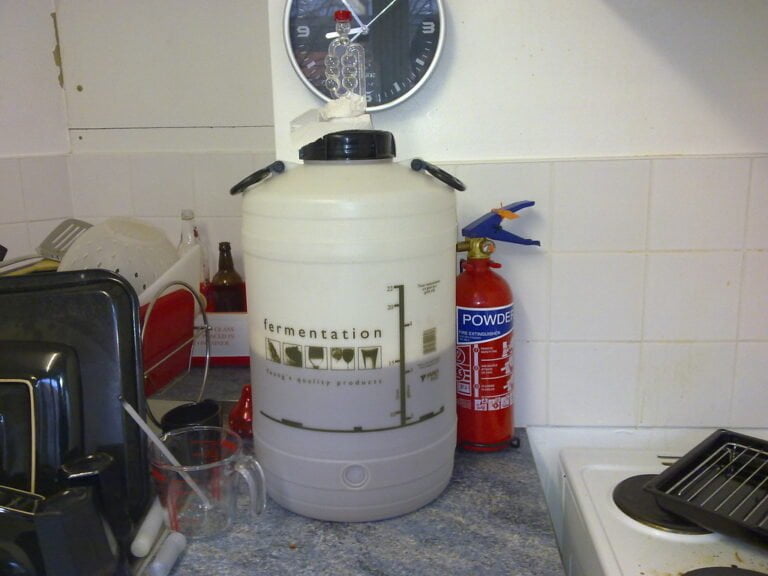 A home brewing fermentation vessel with an airlock on top is placed on a kitchen counter alongside kitchen appliances and a fire extinguisher.