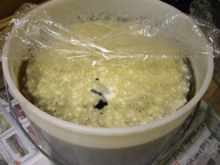 A large plastic container of fermenting beer covered with clear plastic cling film, showing bubbles and foam formation with scattered dark particles on top, resting on a surface covered with newspaper.
