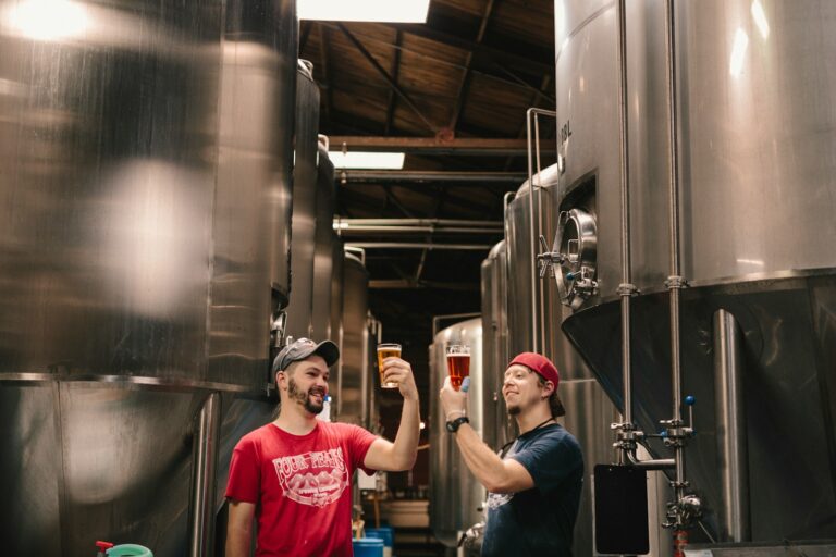Two men smiling and toasting with beer glasses inside a brewery with stainless steel fermentation tanks.