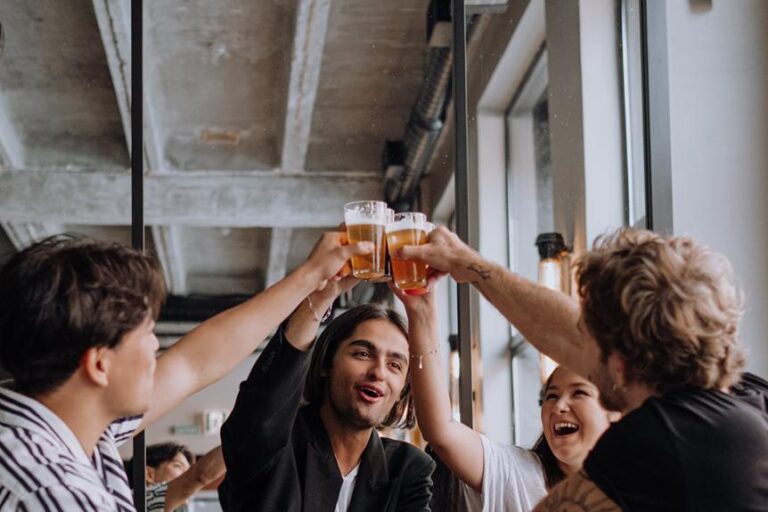 A group of young adults toasting with beer glasses in a casual indoor setting, expressing joy and celebration.