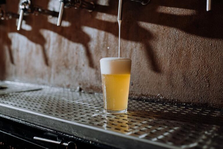 A glass of beer with foam on top is sitting on a metal drip tray, with beer being poured into it from a tap above.
