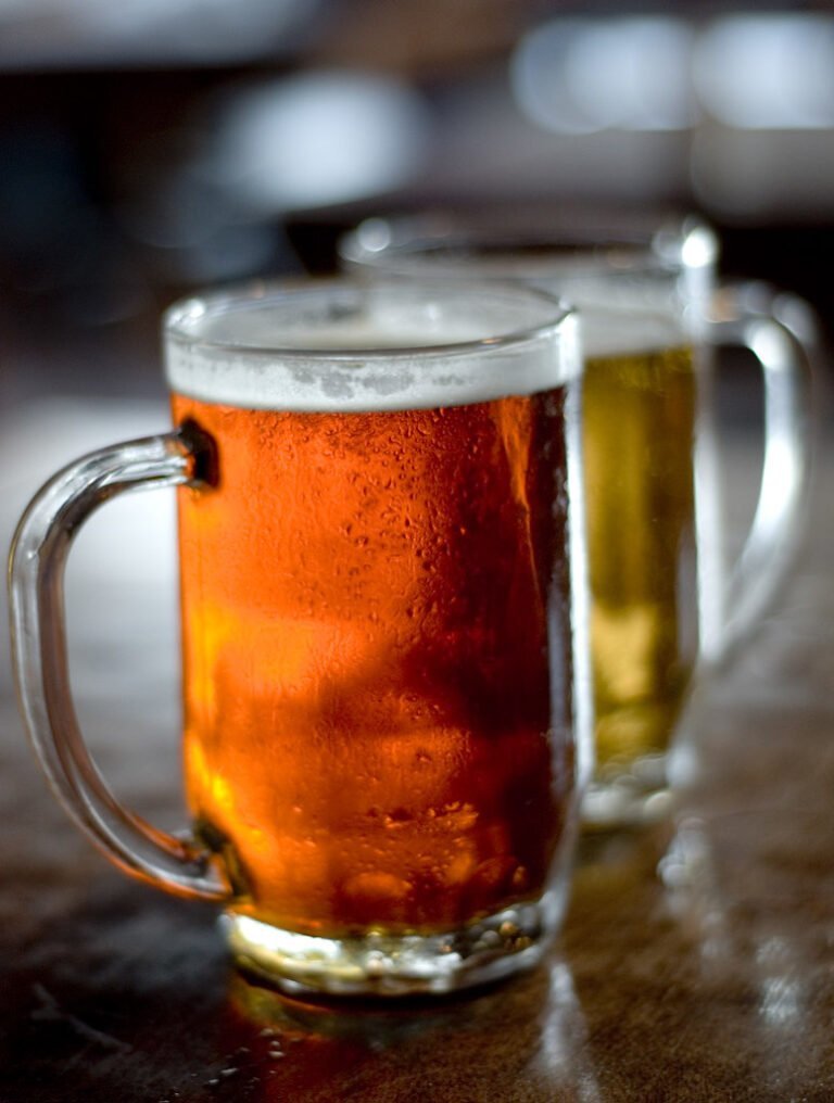 Two frosted beer mugs on a table, one with amber beer in the foreground and the other with pale beer slightly out of focus in the background.