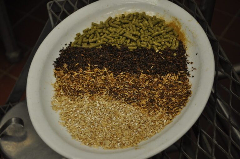 A bowl containing layers of various grains and hops, possibly ingredients for brewing beer, with the colors ranging from pale yellow to dark brown.