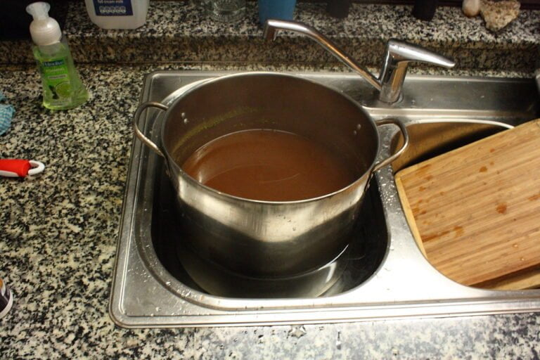 A large pot filled with liquid sits on a stainless steel kitchen sink beside a wooden cutting board and a dish soap dispenser.