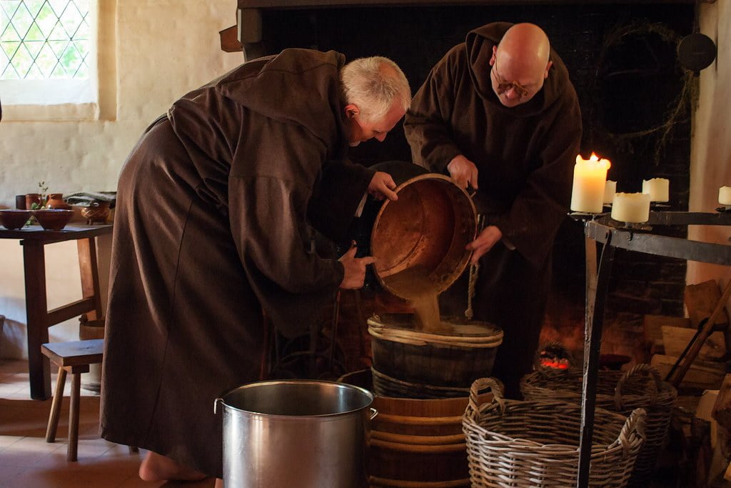 Two individuals in brown monastic robes pour liquid from a large wooden barrel into a metal container, with lit candles and a fireplace nearby, invoking a historical or reenactment scene.