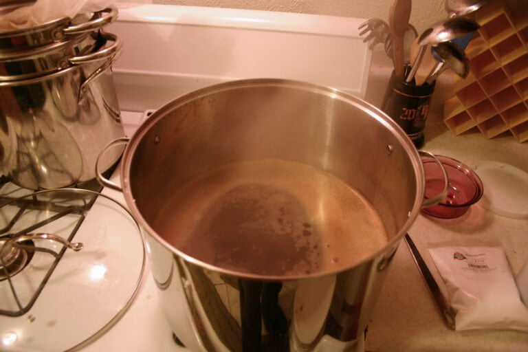 A stainless steel pot with steam rising from it sits on a stovetop next to a stack of pots, cooking utensils, and kitchenware with a bag labeled "Hops" in the foreground.
