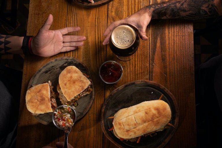 Two people sitting at a wooden table with food and drinks. One person is reaching for a glass of dark beer. On the table are two plates with sandwiches and a side of sauce, with a person's outstretched hand visible on the left.