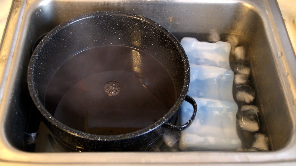 A black frying pan and a blue ice cube tray in a stainless steel kitchen sink filled with water and soap suds.