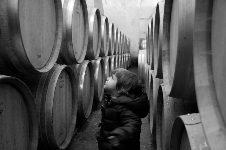 A young child in a black jacket gazes up at large wooden barrels stacked in rows inside a cellar, captured in black and white.