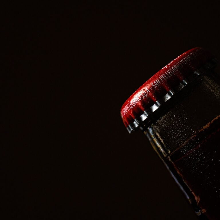 Close-up of the top part of a dark glass bottle with a red cap, featuring condensation against a dark background.