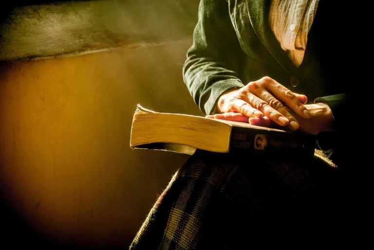 Person holding an open book on their lap, with sunlight illuminating the book and dust particles in the air, creating a peaceful atmosphere.