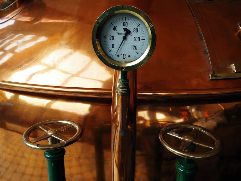 A close-up of a polished copper brewing kettle with a temperature gauge and two valve wheels.