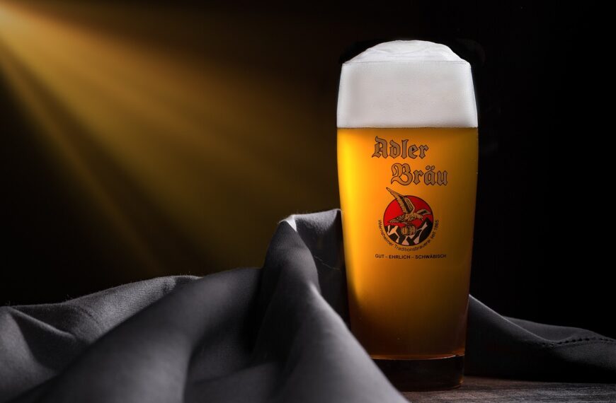 A glass of beer with a logo that reads "Adler Bräu" on it, displayed on a wooden surface with a draped gray cloth, illuminated by a warm, angled light from the left creating a soft glow on the glass and a dark background.