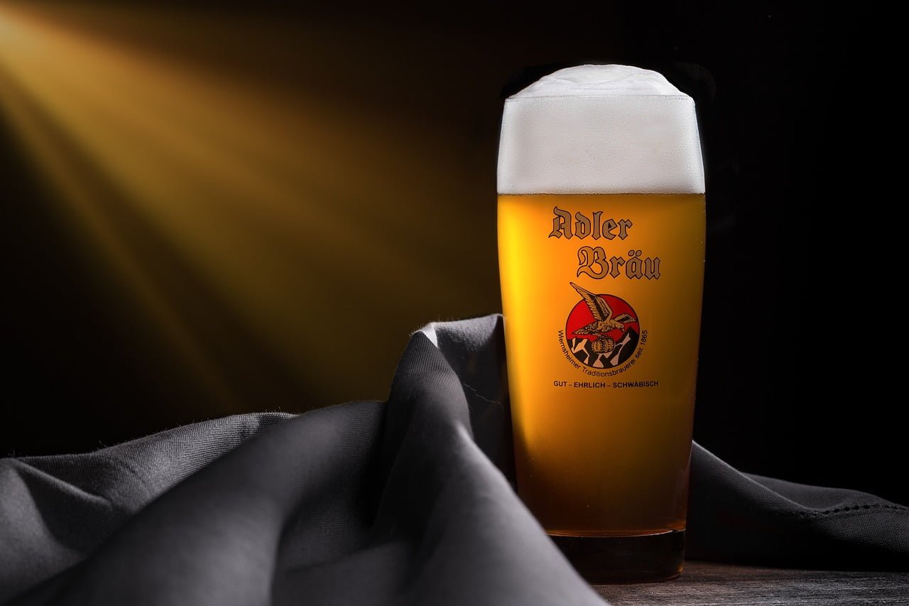 A glass of beer with a logo that reads "Adler Bräu" on it, displayed on a wooden surface with a draped gray cloth, illuminated by a warm, angled light from the left creating a soft glow on the glass and a dark background.